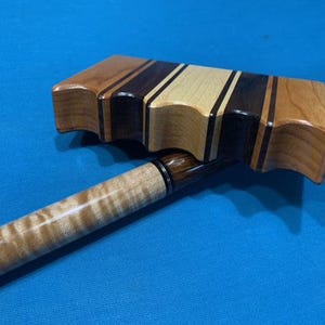 May include: A wooden billiard bridge head featuring a multi-toned striped pattern of light and dark wood grains, resting on a pool cue with a light-colored curly maple shaft and a dark, highly figured wood handle with a black ring accent.