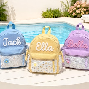 May include: Three corduroy backpacks in blue, yellow, and purple. Each bag features a front pocket with a white floral quilted pattern, a top handle, and a name embroidered in white or pink script: Jack, Ella, and Bella, respectively.