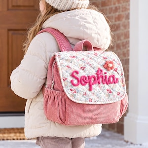 May include: A pink corduroy backpack featuring a floral quilted flap with the name Sophia embroidered in pink yarn and a small fabric flower accent. The bag includes side pockets and a top handle, worn over a beige puffer jacket and knit hat.