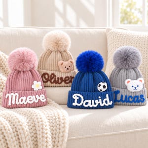 Op de afbeelding: Vier gebreide mutsen met pompons. De roze muts heeft een witte bloem en de naam Maeve. De beige muts heeft een teddybeer-patch en de naam Oliver. De blauwe muts toont een voetbal en de naam David. De grijze muts heeft een beertje en de naam Lucas.