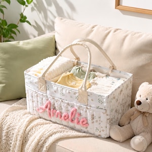 Könnte beinhalten: Ein rechteckiger Aufbewahrungskorb aus Stoff mit weißem Blumenmuster und zwei Henkeln. Der Name Amelie ist in rosa Garn auf der Vorderseite eingestickt. Der Korb ist mit gefalteter Babykleidung in Weiß, Gelb und Hellgrün gefüllt.