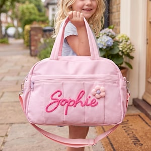 May include: A light pink rectangular bag with two top handles and an adjustable shoulder strap. The front features the name Sophie embroidered in dark pink cursive script and a small, textured, light pink flower applique with a purple center.