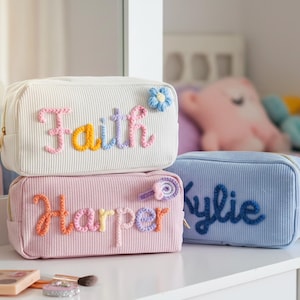 May include: Three personalised corduroy cosmetic bags in cream, pink, and blue. The bags are embroidered with the names "Faith", "Harper", and "Kylie" in colourful thread. Each bag has a zip closure and decorative accents.