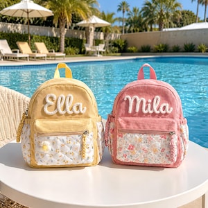 May include: Two corduroy backpacks, one yellow and one pink, featuring white embroidered names Ella and Mila. Each bag has a quilted front pocket with a floral pattern, a top handle, and side pockets, displayed on a white table outdoors.