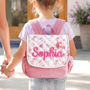 Könnte beinhalten: Ein gesteppter Rucksack mit Blumenmuster auf der Klappe und einfarbigem rosa Kordstoff an den Seiten und Trägern. Der Name Sophia ist in rosa Schreibschrift auf die Frontklappe gestickt, die mit einer kleinen rosa Stoffblume verziert ist.