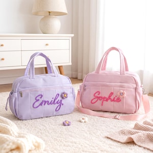 May include: Two pastel-coloured baby bags. One is lavender with "Emily" embroidered on the front, and the other is pink with "Sophie" embroidered. Both bags have a flower accent and top handles.
