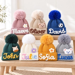 May include: Seven knit beanies with pom-poms, each featuring a unique name and decorative patch. The hats are pink, beige, blue, dark green, white, yellow, and gray, with embroidered names including Maeve, Oliver, David, John, Julie, Sofia, and Lucas.