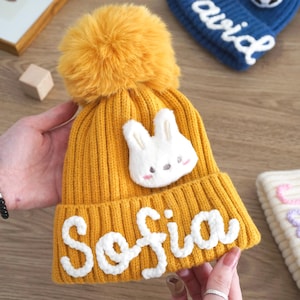 May include: Mustard yellow knit beanie with a fluffy pom-pom and the name "Sofia" in white script. A white plush bunny is sewn on the front. The beanie is a cozy winter accessory.