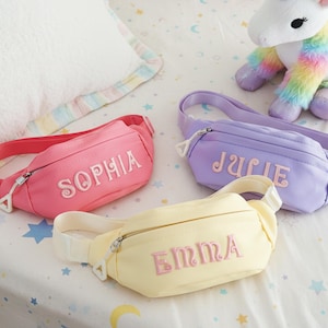 Puede incluir: Tres riñoneras personalizadas en rosa, lavanda y amarillo, cada una bordada con un nombre en un color a juego. Las bolsas tienen cierre de cremallera y correas ajustables. Un unicornio de peluche está en el fondo.