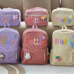 May include: Six corduroy backpacks and lunch bags in pastel colors, each personalized with a name in colorful letters. The bags feature decorative appliques like flowers, a bunny, a lion, and a strawberry. The names are Elise, Bettie, and Molly.