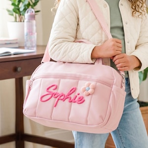 May include: A pale pink quilted duffel bag with the name "Sophie" embroidered in pink script. A pink and blue flower decorates the bag. The bag has a shoulder strap and side pockets.
