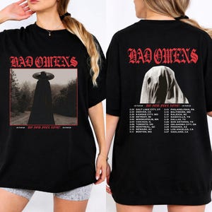 Può includere: T-shirt nero con la scritta rossa "BAD OMENS". Il davanti presenta una figura con cappotto e cappello neri. Il retro ha una figura spettrale e un elenco di date del tour. Un capo a tema musicale.