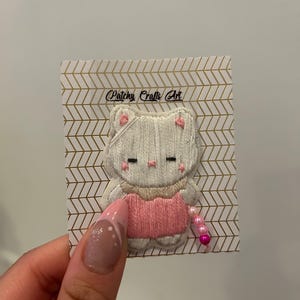 May include: Embroidered cat patch with a pink dress and a string of pink beads. The cat is white with pink accents on the ears and cheeks. The patch is attached to a card with the text "Patchy Crafts Art".