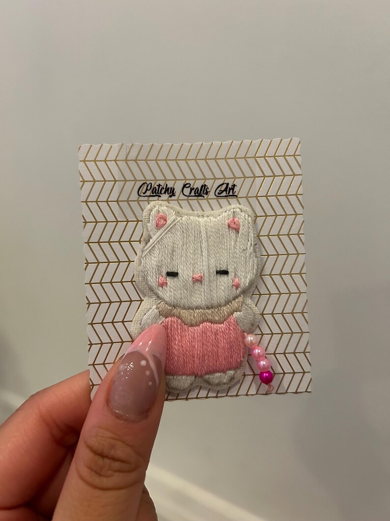 May include: Embroidered cat patch with a pink dress and a string of pink beads. The cat is white with pink accents on the ears and cheeks. The patch is attached to a card with the text "Patchy Crafts Art".