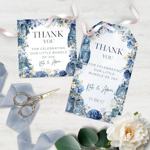 Può includere: Due etichette regalo rettangolari bianche con nastri rosa chiaro, decorate con illustrazioni di giacche di jeans e scarpe da ginnastica blu, fiori blu e il testo: THANK YOU FOR CELEBRATING OUR LITTLE BUNDLE OF JOY. KATE & ADAM. Un'etichetta include SAT 04 OCT.