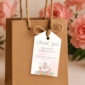 May include: A brown paper gift bag with a tag that reads "Thank You for making my day magical!" The tag has a watercolor design with a fairy and flowers. The bag has a twine handle and bow. Pink flowers are in the background.