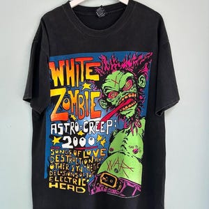 Może przedstawiać: Czarny t-shirt z kolorową grafiką zielonego zombie z czerwonymi oczami i długim językiem. Tekst "WHITE ZOMBIE" i "ASTRO-CREEP 2000" są w kolorze żółtym i pomarańczowym. Koszulka wisi na białym wieszaku.