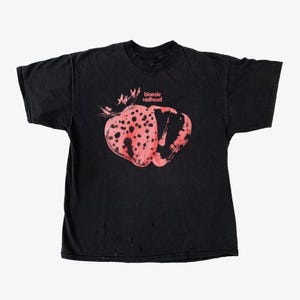 Könnte beinhalten: Schwarzes T-Shirt mit einem rosa und roten Grafikdesign und dem Text "blonde redhead". Das Design ähnelt einer Erdbeere oder einer ähnlichen Frucht mit dunklen Flecken. Das T-Shirt hat einen abgenutzten, Vintage-Look.