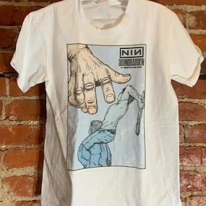 May include: White t-shirt with a graphic depicting a large hand reaching towards a person holding a stick. The graphic includes the text "NIN SOUNDGARDEN" within a rectangular frame.