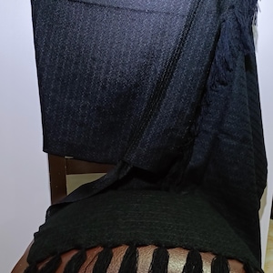 Op de afbeelding: Een zwarte sjaal met een geweven textuur en franjes. De sjaal is over een stoel gedrapeerd en toont de donkere kleur en de gedetailleerde textuur van de stof. De franjes hangen aan de onderkant.