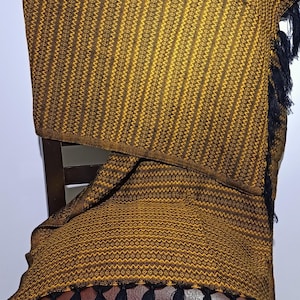 Peut inclure: Un textile tissé avec un motif géométrique répétitif dans des tons or et marron. Le tissu est drapé sur une chaise, avec des franges noires sur les bords. Le motif présente des éléments verticaux et en forme de losange.