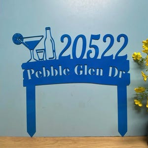 Puede incluir: Un letrero de dirección azul con los números "20522" y las palabras "Pebble Glen Dr" recortadas. El letrero también presenta un diseño de cóctel, botella y vaso. El letrero está hecho de un material sólido y tiene una estaca para una fácil colocación.