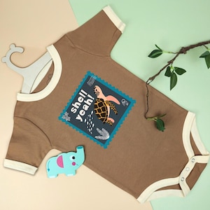 May include: Brown baby bodysuit with cream trim and a blue and teal graphic of a turtle with the words "shell yeah!". A light blue elephant-shaped wooden toy is next to the bodysuit.