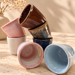 May include: A collection of six small ceramic cups with varied reactive glazes in shades of brown, blue, pink, and white. The cups are arranged in a stacked formation, showcasing smooth, glossy interiors and textured, speckled, or mottled exterior finishes.