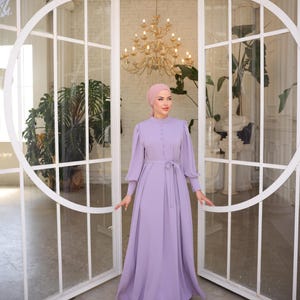 May include: A long, lavender dress with long sleeves and a matching belt. The dress features a high neckline and button details. The person is wearing a pink head covering and silver embellished shoes. The background includes large glass doors and a chandelier.