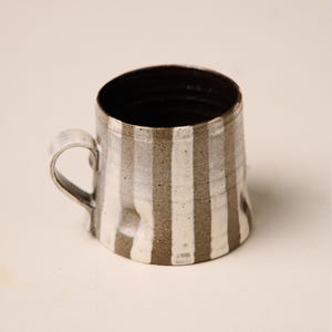 May include: A handmade ceramic mug with a unique, textured design. The mug features vertical stripes in alternating shades of white and brown. It has a curved handle and a dark interior, perfect for coffee or tea.