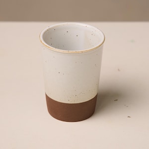 Może przedstawiać: Ręcznie robiony ceramiczny kubek o dwutonowym wzorze. Górna część jest nakrapiana na biało, a podstawa jest w kolorze głębokiego brązu. Kubek ma cylindryczny kształt i lekko teksturowaną powierzchnię.