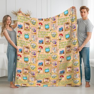 May include: A peach-colored blanket featuring a repeating pattern of postage stamp-style frames containing illustrations of characters from Toy Story, including Woody, Buzz Lightyear, and Jessie, with the text YOU'VE GOT A FRIEND IN ME.
