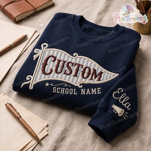 May include: A navy blue crewneck sweatshirt featuring an embroidered pennant design on the chest with the word CUSTOM in dark red letters and the text SCHOOL NAME below. The sleeve includes the name Ella embroidered above a megaphone icon.