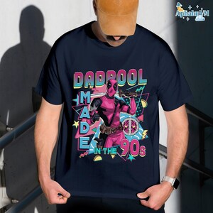 A person wearing a black t-shirt with a graphic design featuring a character from the Marvel Cinematic Universe, specifically the character Deadpool. The design includes the text "DAD POOL MADE IN THE 90'S" and a peace sign.