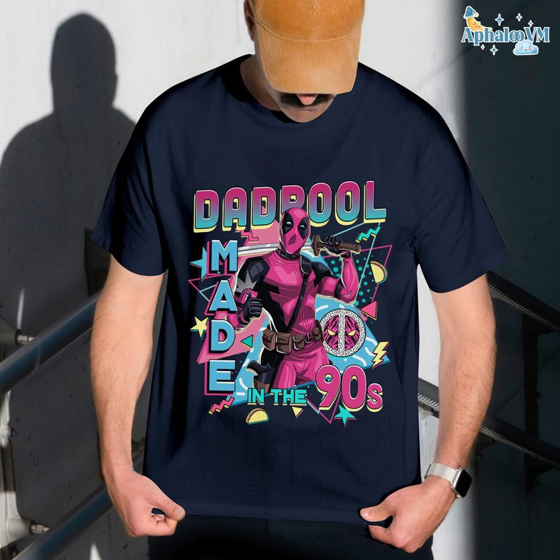 A person wearing a black t-shirt with a graphic design featuring a character from the Marvel Cinematic Universe, specifically the character Deadpool. The design includes the text "DAD POOL MADE IN THE 90'S" and a peace sign.