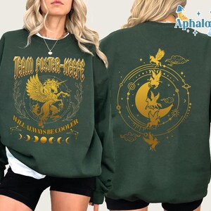 May include: Dark green sweatshirt with gold graphic design. The front features a winged unicorn, text that reads "Team Foster-Keefe" and "Will Always Be Cooler". The back has a gold crescent moon and fairy design.