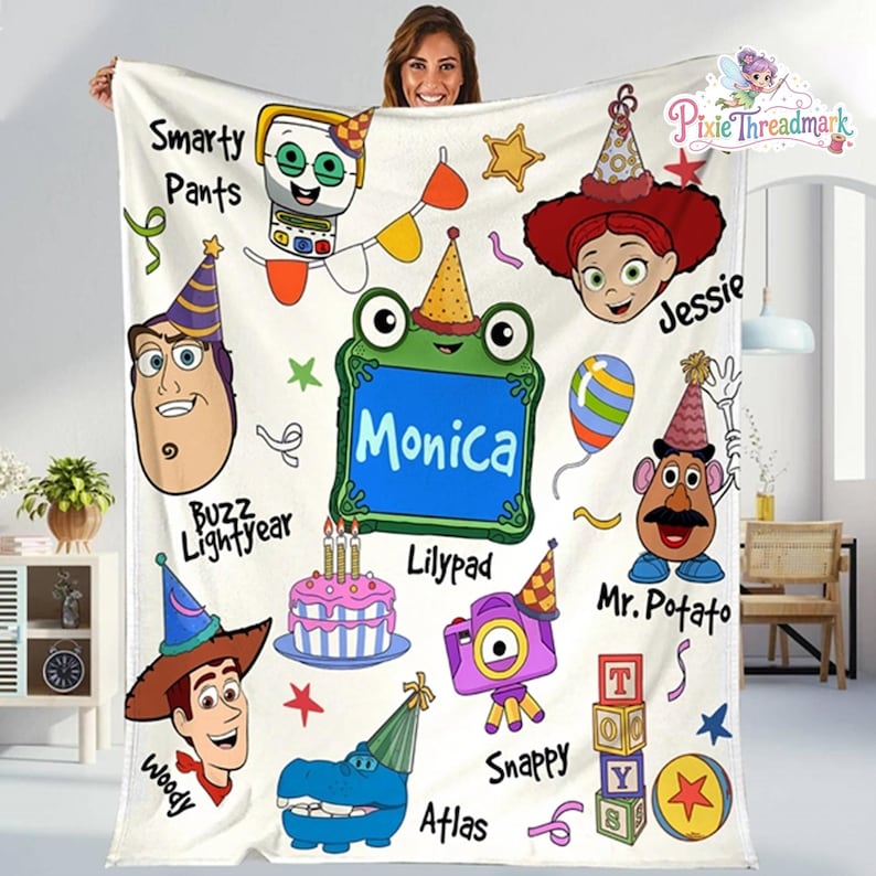 May include: A white fleece blanket featuring a colorful birthday-themed print with cartoon characters including Buzz Lightyear, Woody, Jessie, and Mr. Potato Head. The design includes a central green frog frame with the name Monica, a cake, and text.