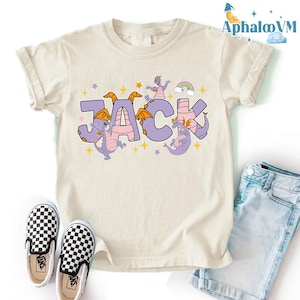 May include: Cream-colored t-shirt featuring the name "JACK" in large, purple letters with pink accents. Dragons are incorporated into the design, along with stars and a rainbow. Checkered slip-on shoes and denim shorts are also visible.