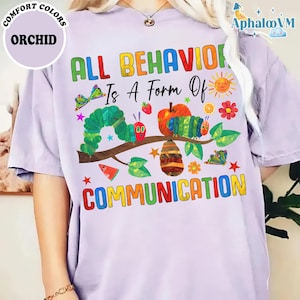 May include: A light purple t-shirt with the text "ALL BEHAVIOR IS A FORM OF COMMUNICATION" in colorful letters. The shirt features a whimsical illustration of a caterpillar, fruits, and flowers.
