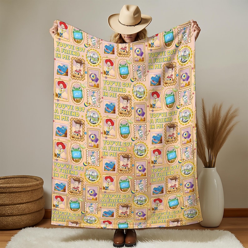 May include: A light peach-colored throw blanket featuring a repeating pattern of Toy Story characters including Woody, Buzz Lightyear, and Rex, framed in yellow and orange borders with the text "YOU'VE GOT A FRIEND IN ME" printed in green.