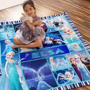 May include: A blue and white patterned blanket featuring images of Elsa from the movie Frozen. The blanket has a blue border with white geometric designs and the name "ELSA" in large letters. A young girl is sitting on the blanket.