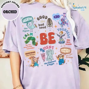 Op de afbeelding: Orchidee-kleurig T-shirt met een grafisch ontwerp. Het ontwerp bevat het woord "BE" in het rood, omringd door stripfiguren en positieve woorden als "Creatief", "Vriendelijk" en "Lief". Het shirt heeft ook de woorden "Comfort Colors" en "Orchid" erop gedrukt.
