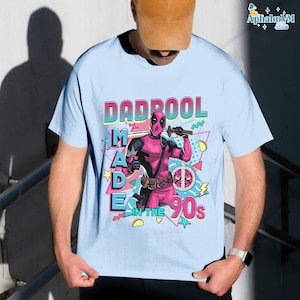 A person wearing a t-shirt with a graphic design featuring a character from the Marvel Cinematic Universe, specifically the character Deadpool. The design includes the text "DAD BOOL MADE IN THE 90'S" and a peace sign.