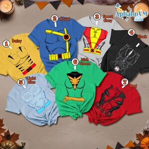 May include: A collection of seven t-shirts in various colors, each featuring a superhero costume design. Colors include yellow, blue, gray, black, light blue, green, and red. The shirts are displayed on a wooden surface.