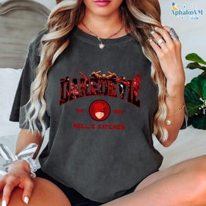 May include: Dark gray t-shirt featuring the word "DAREDEVIL" in red, with a graphic of characters and the text "EST. 1964 HELL'S KITCHEN". The design includes a red circle with a mask. The shirt has short sleeves.