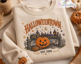 May include: A beige crewneck sweatshirt featuring an embroidered design of a haunted house, a large jack-o'-lantern, and a broomstick. The text HALLOWEENTOWN and EST. 1998 are stitched in orange. A small jack-o'-lantern is embroidered on the sleeve cuff.