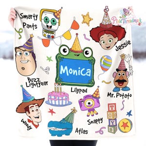 May include: A white fleece blanket featuring a colorful Toy Story character print with party hats, balloons, and confetti. Characters include Woody, Buzz Lightyear, Jessie, and Mr. Potato Head, alongside a frog named Monica, a camera, and a cake.