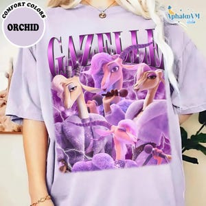 A person wearing a purple t-shirt with a graphic design featuring purple and pink cartoon-like characters, including a unicorn and a bird, along with the text "Gazelle" and "Orchid".