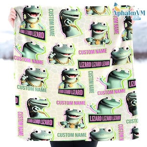 May include: A light-colored blanket featuring a repeating pattern of cartoon lizards. Each lizard is wearing a gold chain and has the text "Lizard Lizard Lizard" and "Custom Name" on the blanket. The blanket is held up by a person.