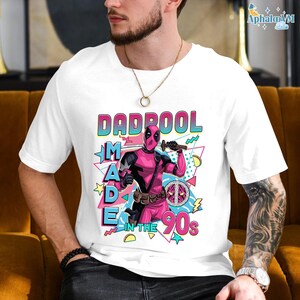 A white t-shirt with a graphic design featuring a character from the Marvel Cinematic Universe, specifically the character Deadpool. The design includes the text "DAD POOL MADE IN THE 90'S" and includes various colorful elements and symbols associated with the character.